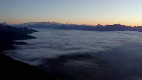 Flying-high-over-fog-at-dusk,-the-Alps-in-the-background-glowing-orange---Switzerland
