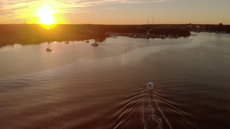 Aerial-view-of-a-motorboat-entering-a-beautiful-small-town-harbor-on-a-calm-river-under-a-colorful-sunset
