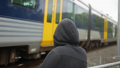Slow-motion-of-a-blonde-man-wearing-a-black-hoodie-standing-and-looking-at-a-train-passing-by-on-railroads,-in-a-cloudy-day