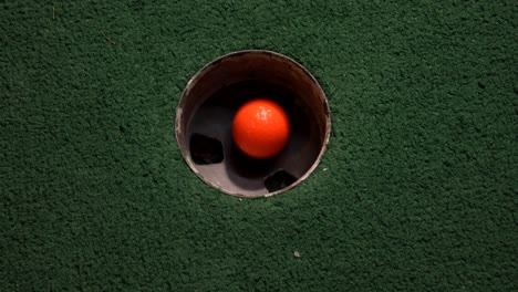 A-close-up-birds-eye-view-of-an-orange-mini-golf-ball-falls-into-the-golf-hole-and-bounces-in-the-pocket-on-a-course