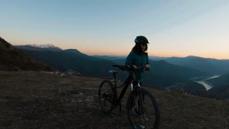 Chica-Empujando-Su-Bicicleta-En-Una-Colina-De-Montaña-Al-Atardecer-Con-Un-Hermoso-Lago-De-Cañón-En-El-Fondo