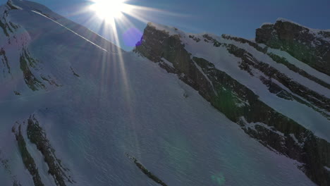 Aerial-view-pulling-back-from-a-shadowy-mountain-ridge-valley-with-sun-in-shot-creating-light-flare-in-the-French-Alps-in-winter
