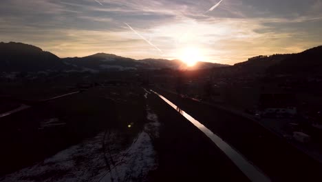 Wunderschöne,-Stimmungsvolle-Winterlandschaft-In-Der-Schweiz-Mit-Einem-Reflektierenden-Fluss,-Autos-Und-Einem-Dorf-Bei-Sonnenuntergang