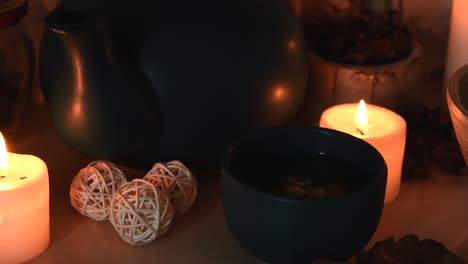 Relaxing-background-detail-shot-of-an-herbal-tea-on-a-green-cup,-with-steam-coming-out,-near-candles-with-flickering-flames,-a-tea-pot-and-some-herbs