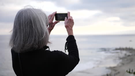 Una-Anciana-De-Vacaciones-Usando-Su-Teléfono-Para-Tomar-Una-Foto-En-La-Playa-Sobre-El-Océano-Al-Atardecer-En-Cámara-Lenta