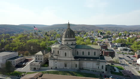 Luftdrohnenansicht-Der-Kathedrale-Des-Allerheiligsten-In-Altoona,-Pennsylvania,-Mit-Der-Amerikanischen-Flagge-Dahinter