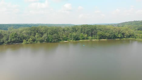 Drone-flight-right-over-forest-lake-in-Illinois-with-lush-green-trees-and-blue-cloudy-sky-on-a-sunny-day-as-people-float-on-the-water-in-a-kayak-4k