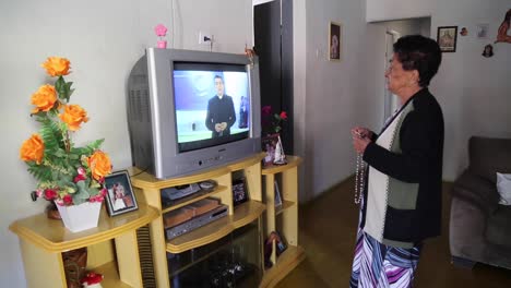 A-Woman-holds-her-rosary-while-praying-in-front-of-a-tv-set-listening-to-the-priest