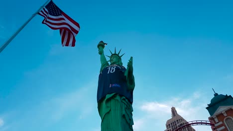 Panning-shot-of-Statue-of-Liberty-wearing-basketball-shirt-and-US-Flag-waving-in-the-wind-outside-New-York-New-York-Hotel-and-Casino-in-Las-Vegas,-Nevada,-USA
