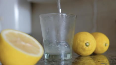 Gießen-Sie-Limonade-Und-Eiswürfel-In-Ein-Glas-Zwischen-Zitronen,-Die-Auf-Die-Arbeitsplatte-Spritzen