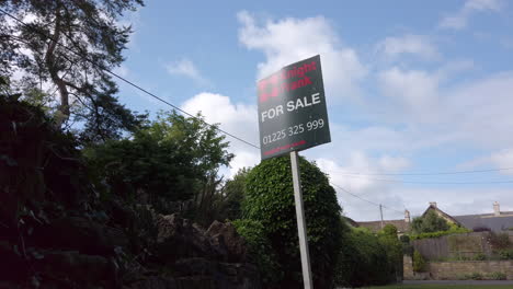 Low-Angle-Static-Shot-of-British-‘For-Sale’-Real-Estate-Sign-on-Sunny-Summer’s-Day-with-Foliage-Blowing-in-the-Breeze-in-the-Background