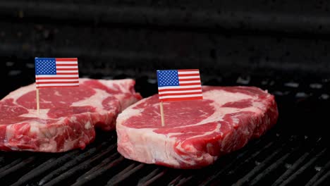 Two-juicy-rib-eye-steaks-sitting-on-the-grill-and-cooking-with-two-tiny-American-flags-tooth-picked-into-them