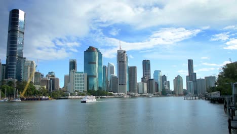 Klassischer-Blick-Auf-Das-Zentrum-Von-Brisbane-Von-Der-Anderen-Seite-Des-Flusses,-Wobei-Die-Sonne-Die-Modernen-Hochhäuser-Der-Stadt-Betont,-Während-Eine-Flussfähre-Durch-Den-Vordergrund-Fährt