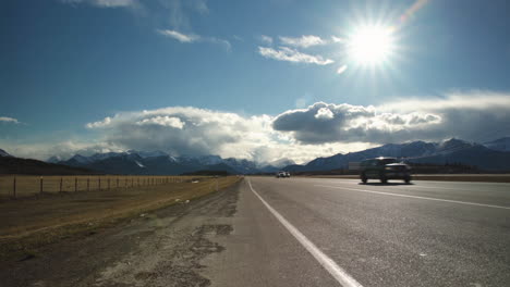 Vehicles-driving-down-highway-with-beautiful-mountains-and-clouds-in-the-background-as-the-sun-shines-overhead