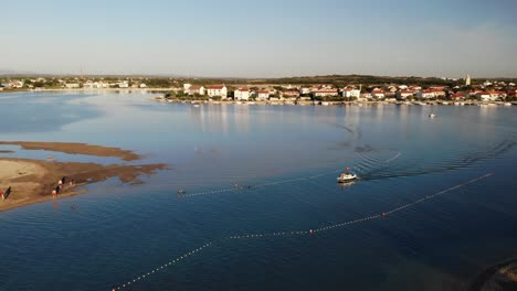 Aerial-panorama-of-a-Nin-town-with-lagoon-and-following-boat-approaching-sandy-beach-in-sunset
