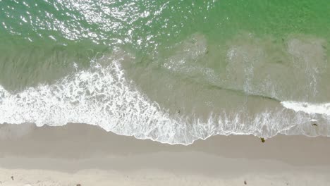 Low-altitude-birds-eye-view-over-waves-crashing-on-a-sandy-beach-during-a-bright-and-sunny-afternoon