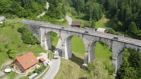 Aerial-high-angle-follow-shot-of-truck-driving-on-the-country-road-and-passing-below-railway-viaduct-in-Slovenia