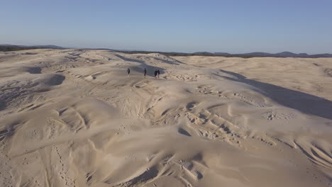 People-walking-across-sand-dunes-in-Newcastle,-Australia,-low-aerial-follow