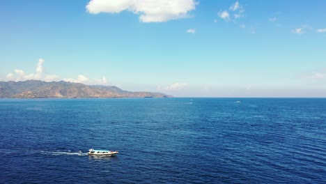 Barco-Turístico-Con-Turistas-Navegando-A-Través-Del-Mar-Azul-Profundo,-Acercándose-A-La-Bahía-De-La-Isla-Tropical-Sobre-Un-Fondo-De-Cielo-Brillante-Con-Nubes-Blancas-En-Bali