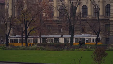 Gelbe-Straßenbahn-Fährt-Vor-Dem-Nationalen-Ethnografischen-Museum-In-Budapest,-Ungarn,-In-Der-Nähe-Von-Grünem-Gras-Und-Kahlen-Bäumen-Vorbei---Panoramaaufnahme