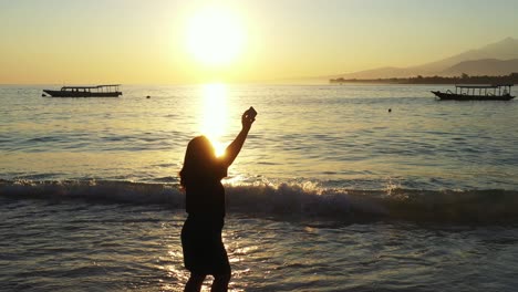 Silhouette-of-young-woman-taking-selfie-pictures-on-calm-beach-at-sunset-golden-hour-with-yellow-sky-reflecting-on-sea-surface