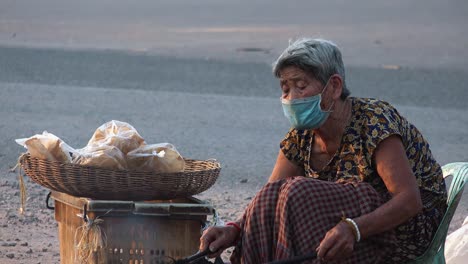 Medium-Shot-of-Lady-With-a-Face-Mask-Using-Long-Sticks-Cooking-Flat-Bread-Over-an-Smoky-Fire-on-the-Road-Side-as-Motorcycles-Drive-Past-in-the-Background
