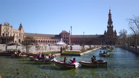 Tourists-row-rental-boats-in-canal-of-Plaza-de-Espana,-Seville,-Slowmo-pan-left