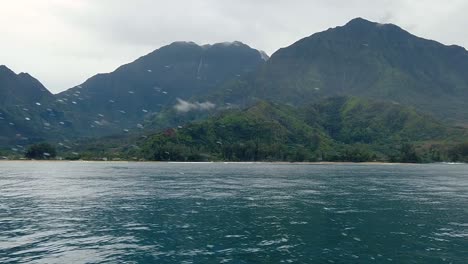 HD-120fps-Hawaii-Kauai-Boating-on-the-ocean-floating-right-to-left-green-hills-and-mountain-with-waterfall-in-cloudy-distance-with-boat-spray-in-foreground