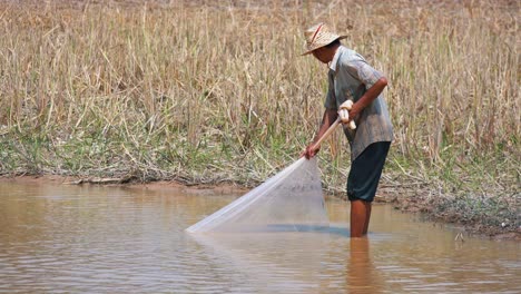 Wide-Exterior-Shot-of-Fisherman-in-Straw-Hat-Carefully-Drawing-The-Net-In,-in-the-River-Close-to-the-Grassy-Shoreline-in-the-Daytime