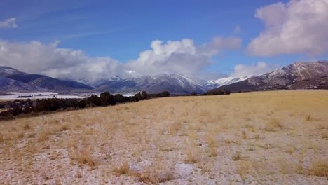 Aerial-view-of-pasture-landscape-with-snow-covered-mountains,-clouds-and-blue-sky-in-the-distance---dolly-forward