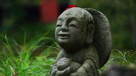 Eine-Kleine-Jizo-Statue-Mit-Einem-Roten-Blatt-Auf-Dem-Kopf,-Die-Im-üppigen-Grünen-Gras-Steht