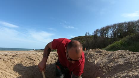 A-Man-Digging-A-Hole-On-The-Beach-Sand-In-Brittas-Bay-Using-A-Shovel-In-County-Wicklow,-Ireland-On-A-Sunny-Day