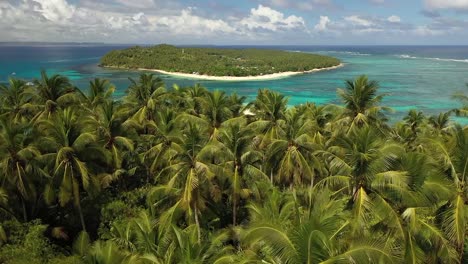 Aerial-shot-flying-right-over-the-top-of-the-palm-trees-in-the-jungle,-revealing-a-beautiful-and-quiet-white-sand-beach-with-a-small-island-nearby-in-the-secluded-paradise-with-a-few-small-boats