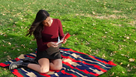 Eine-Schöne-Hispanische-Studentin,-Die-Ein-Buch-Liest-Und-Sich-Draußen-Im-Park-Mit-Herbstblättern-Und-Grünem-Gras-Entspannt
