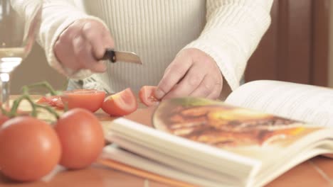 Manos-Cortando-Tomates-De-Vid-Frescos-En-La-Cocina-Con-Un-Libro-De-Recetas-De-Tiro-Medio