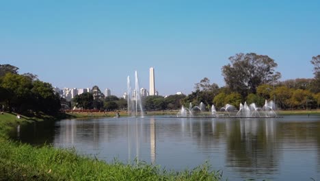 Panorámica-Del-Lago-Y-Las-Fuentes-Del-Parque-Ibirapuera,-Con-Paisaje-Urbano-Y-Obelisco-En-La-Parte-Posterior