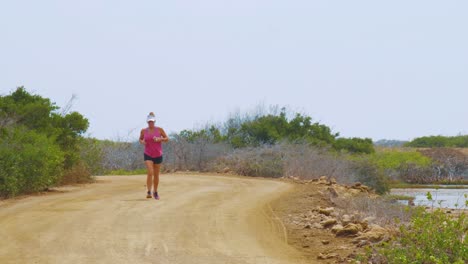 An-Active-Woman-With-Sun-Visor-Cap-Running-On-The-Dirt-Road-In-Kralendijk,-Bonaire---midshot