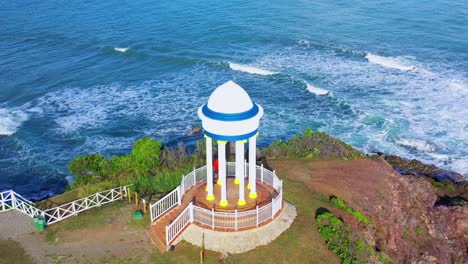 Colorful-structure-at-rocky-seaside-summit-with-green-ocean-waves-crashing-onshore,-Puerto-Plata,-Dominican-Republic,-above-circle-aerial