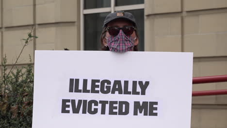 A-protestor-wearing-a-face-mask-on-a-housing-demonstration-opposing-eviction-during-the-Coronavirus-pandemic,-stands-holds-a-white-cardboard-placard-that-says,-“Illegally-evicted-me”