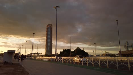 Cars-drive-by-people-and-Torre-Pelli-in-golden-sunset-landscape,-Seville,-Spain