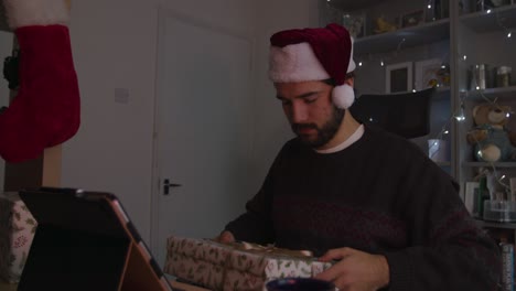 Male-opening-Christmas-gift-on-a-videocall-while-wearing-a-Christmas-hat