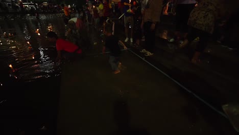 Ein-Thailändischer-Junge-Vergnügt-Sich-Im-Kanalwasser-Der-Stadt,-Während-Andere-Menschen-In-Masken-Loy-Krathongs-Schwimmen-Lassen-Und-Nachts-Während-Der-Loi-Krathong-Feierlichkeiten-In-Korat,-Thailand,-Beten