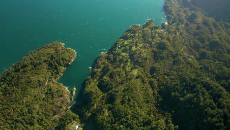 Aerial-view-looking-down-at-the-edge-of-Reloncavi-estuary,-Cochamo,-showing-bright-blue-water-and-green-forests