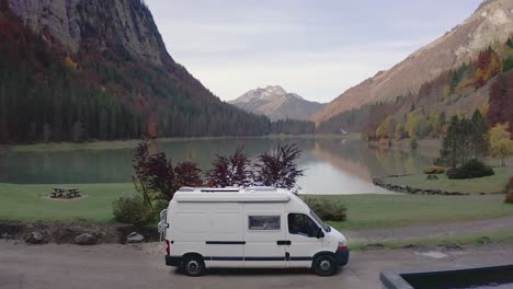 A-white-camper-van-is-slowly-driving-on-a-dirt-road-with-a-gorgeous-mountain-lake-view-in-the-background,-lac-de-Montriond,-French-Alps