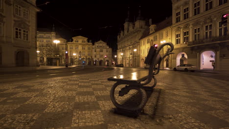 A-bench-on-the-empty-Malostranské-square-with-a-tram-stop-and-rails-in-the-historical-centre-of-Prague,-Czechia,-at-night-lit-by-street-lights,-during-a-Covid-19-lockdown-with-no-people-anywhere