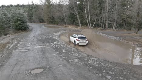 A-single-white-Subaru-Crosstrek-drives-into-a-deep-mud-puddle-splashing-dirty-water,-slow-motion-aerial,-illustrative-editorial