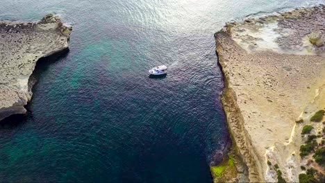 An-early-morning-Birds-Eye-View-of-a-small-boat-turning-around-at-St-Peters-Pool-Malta-displaying-the-golden-sandstone-rocks-and-the-crystal-clear-blue-water-of-the-Medaterainan