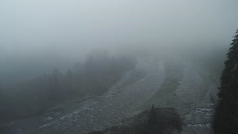 Horror-like-scenery-with-thick-mist-and-dark-trees-next-to-rocky-river,-aerial