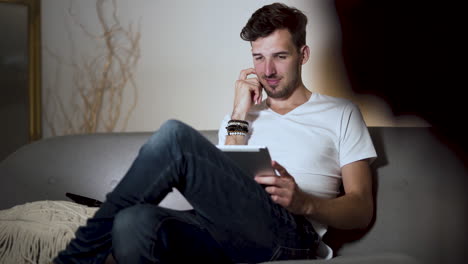 A-young-man-with-a-stubble-and-a-modern-hairstyle,-wearing-a-white-t-shirt-and-jeans,-sitting-at-home-on-a-sofa,-legs-crossed,-laughing-while-browsing-his-tablet,-static-4k