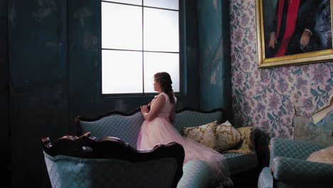 Young-girl-in-pink-dress-looks-over-her-shoulder-from-window-seated-on-blue-couches